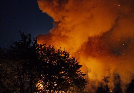 Feuerschein vor Nachthimmel im Vordergrund Umrisse von Laubbäumen.