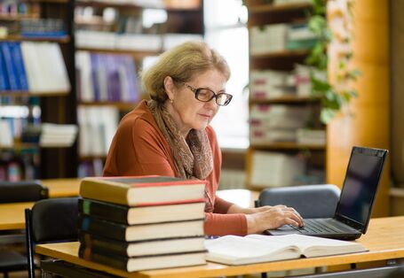 Eine Frau sitzt mit einem Laptop in einer Bibliothek und liest ein Buch.