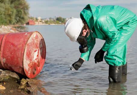 A red barrel lies in a river and is examined by a person in a green protective suit.