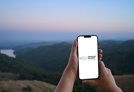 Ein Smartphone wird an einem Fluss benutzt. Auf dem Bildschirm ist zu lesen: "Gewässerkompass Deutschland".