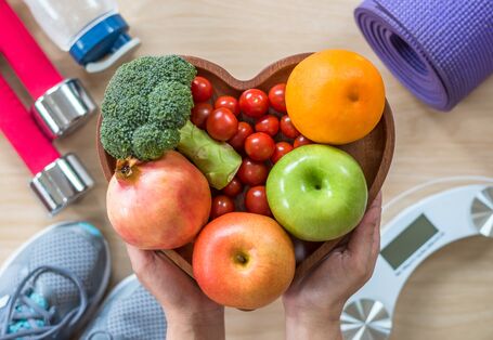 Photo depicting a healthy lifestyle: nutrition with fruit and vegetables in a heart-shaped bowl, physical fitness with sneakers, yoga mat, dumbbells, water bottle, and scale.
