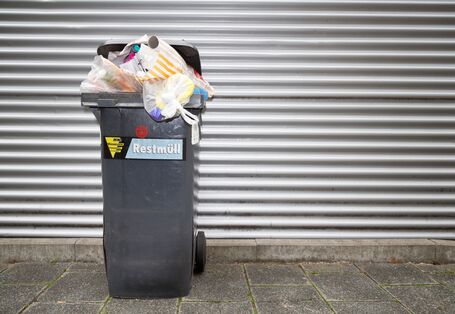 Eine überquellende Abfalltonne vor einem silberfarbenen Tor.