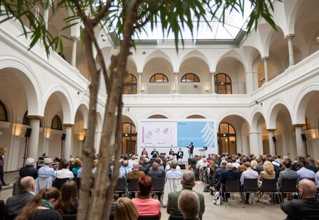 Das Bild wurde auf der Fachkonferenz 2017 zum Thema Vulnerabilität Deutschlands gegenüber dem Klimawandel aufegnommen. Man sieht einen hellen, historischen mit einem Glasdach überdachten Innenhof. Es findet gerade eine Podiumsdiskussion statt. Viele Personen sitzen in Reihen vor der Bühne und hören gespannt zu. Außerdem ist eine PowerPoint erkennbar.  