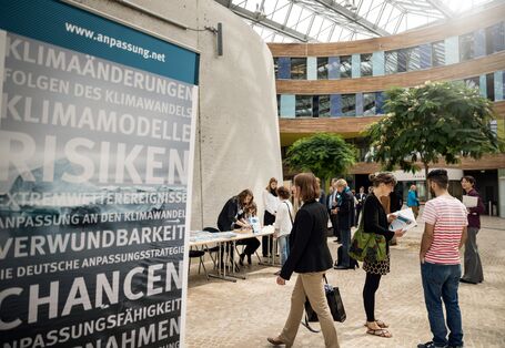 Sie sehen ein Bild vom Nationaler Dialog 2017 Gemeinsam sind wir klimaresilient – Wie Beteiligung in Städten und Regionen gelingt. Das Bild wurde im Umweltbundesamt in Dessau aufgenommen. Auf der rechten Seite sehen Sie ein Aufsteller zum Thema Klimarisiken und Anpassung. Auf der rechten Seite des Bildes sind Menschen auf dem Weg in den Hörsaal in Dessau.