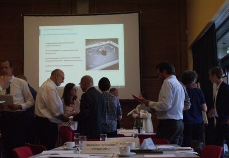 Ein Bild von dem Stakeholderdialog 2019: Katastrophenvorsorge bei Wasserengpässen kooperativ gestalten. Man sieht im Hintergund die PowerPoint Präsentation auf der das Planspiel erklärt ist, welches im Vodergrund abläuft. Personen stehen in kleineren Gruppen zusammen und diskutieren miteinander. 