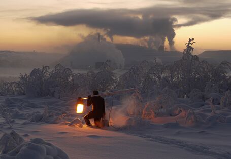 Mann mit leuchtender Laterne in einer eisigen Schneelandschaft. Im Hintergrund rauchende Industrieschornsteine.