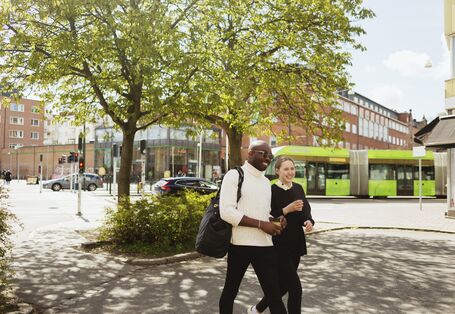 Zwei Menschen laufen lächelnd durch das Bild. Im Hintergrund eine grüne Straßenbahn.