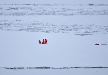Eine Gruppe von Forschern in orangenen Kälteschutzanzügen steht auf einer großen Eisfläche. Rechts von ihnen, am Bildrand steht ihre Ausrüstung.