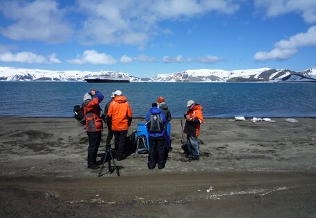 Reisende stehen im Vordergrund auf Land. Im Hintergrund sieht man antarktisches Meer und ein großes Schiff vor beschneiten Hügeln. 