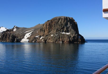 Blick von einem Schiff auf einen schroffen Berg, der aus der arktischen See ragt. 