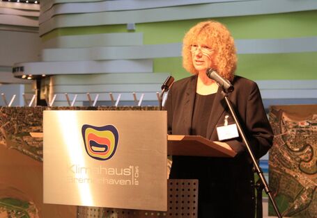 Eine Frau steht an einem silbernen Pult und spricht ins Mikrofon. 