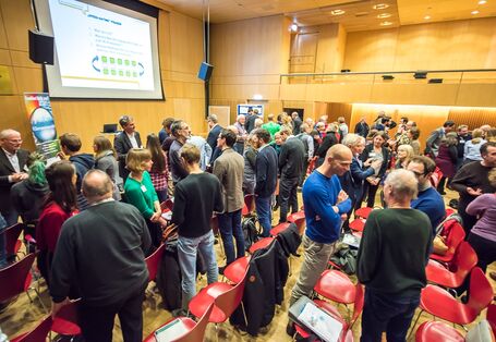 Auf dem Bild ist ein großer Saal zu sehen. Hinter der Bühne ist eine Präsentation an die Wand geworfen. Die Personen im Publikum haben sich alle von ihren Plätzen erhoben und sind in verschiedene Richtungen gedreht. Sie tauschen sich rege miteinander aus.