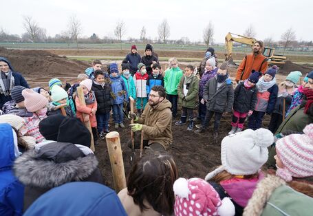 Das Bild zeigt einen Mann, der auf dem Boden hockt und einen kleinen Baum zwischen den Händen hält. Um ihn herum stehen im Kreis viele Kinder und verfolgen die Pflanzmaßnahme.