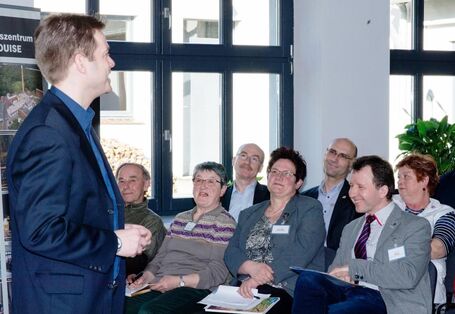 Ein Mann im Anzug steht vor einigen Personen im Publikum und spricht. Er hat die Hänge zusammen gefaltet und schaut geradeaus. Im Publikum sitzen 7 Personen in zwei Stuhlreihen hintereinander und hören zu. Sie tragen Namensschilder