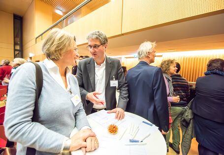 Im Vordergrund stehen zwei Personen an einem Stehtisch. Der Mann erklärt der Frau etwas und gestikuliert mit der Hand. Die Frau schaut ihn gespannt an. Im Hintergrund sind weitere Personen zu sehen, die stehen oder gehen.