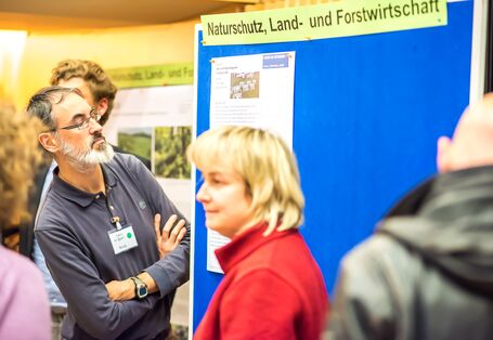 Auf dem Bild ist eine blaue Informationstafel zu sehen. Auf dieser ist ein Text und die große Überschrift: Naturschutz, Land- und Forstwirtschaft. Ein Teilnehmer liest den Text mit dem Kinn nach vorne geschoben und verschränkten Armen durch.