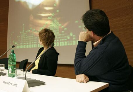 Zwei Podiumsdiskutanten blicken auf eine Leinwand hinter sich, auf der ein Video gezeigt wird.
