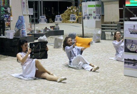 Drei Tänzerinnen sitzen mit überschlagenen Beinen auf dem Boden und halten Plastikfolien wie zeitungen vor ihre Gesichter