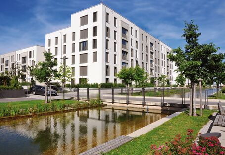 Das Bild zeigt einen künstlichen Wasserlauf, der in einem neu entwickelten Quartier an einem Geschosswohnungsbau vorbeifließt. Eine Straße quert den Wasserlauf. Der Wasserlauf ist von einem schmalen Grünstreifen mit Rasen, jungen Bäumen und Blumenbeeten eingefasst. Bänke bieten Sitzmöglichkeiten.