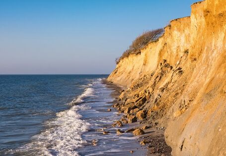 The picture shows a light-brown cliffscape near Ahrenshoop on the Baltic Sea coast. At the foot of the cliff, you can see the sea and the spray of a wave. In one coastal section, you can see loose material and rock fragments which have been shifted from the cliff top to the bottom of the cliff.