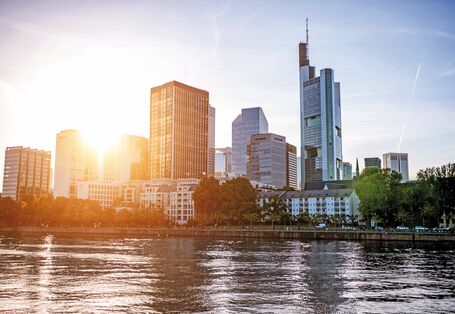 Das Bild zeigt einen Ausschnitt der Skyline von Frankfurt am Main, beschienen von einer tief stehenden Sonne. Im Vordergrund ist der Fluss zu sehen.
