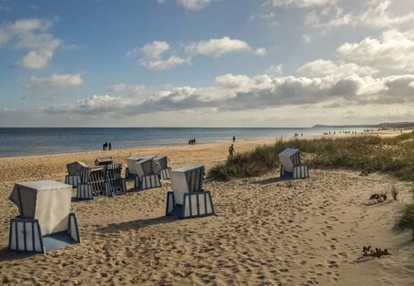 Das Bild zeigt einen Sandstrand an der Ostsee mit Dünen und Strandkörben. Die der oder dem Betrachtenden zugewandten Strandkörbe sind versperrt. Am Horizont ist das Meer zu sehen. An der Wasserlinie sind einzelne Personen und Personengruppen zu erkennen, sonst ist der Stand leer. Der Himmel ist in Teilen bewölkt.