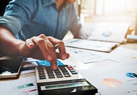 The picture shows a man sitting at a desk holding a ballpoint pen between his fingers while operating a pocket calculator. There are several documents on the desk; it is just possible to make out the fuzzy image of some charts. 