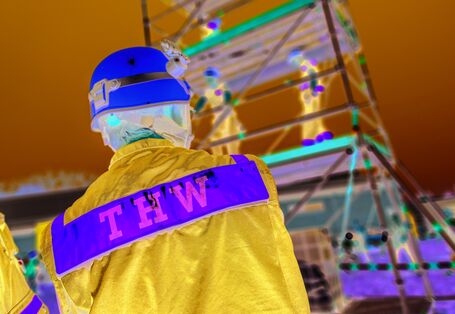 Das Bild zeigt einen Mitarbeiter des Technischen Hilfswerks in Uniform und mit Helm von hinten. Im Hintergrund ist ein Gerüst zu erkennen, auf dem weitere Mitarbeitende des THW stehen.