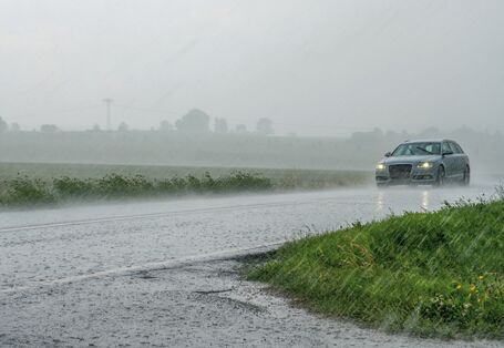 Das Bild zeigt eine mit Wasser bedeckte Straße, auf der ein Auto fährt. Dieses hat Scheinwerfer und Scheibenwischer angeschaltet. Es regnet stark.