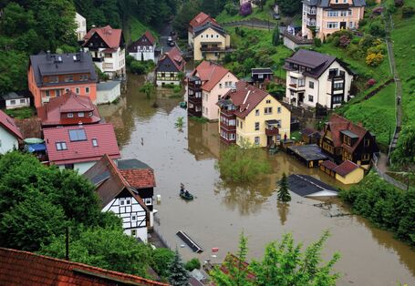 The picture shows a village – situated on the slope of a hill – inundated by floodwater. Some of the lower buildings and some lower floors of houses have been flooded.