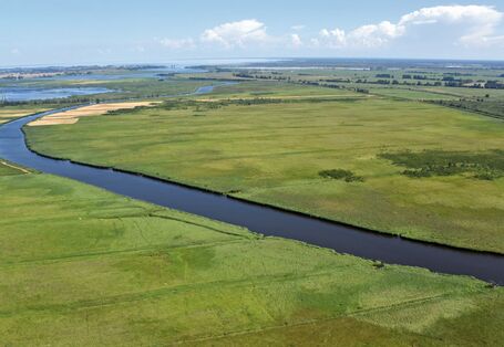 Das Bild zeigt einen Fluss, der durch eine flache Landschaft aus Wiesen und Landwirtschaftsflächen fließt. Im Hintergrund ist das Meer zu sehen.