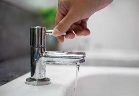 The picture shows a hand on the handle of a tap, with a weak water jet pouring into a wash-hand basin.
