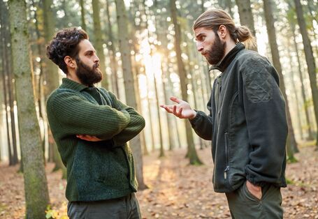 The picture shows two young foresters having a discussion in a sparse forest. The man on the left-hand side of the picture stands with his arms crossed while the other man is gesticulating with his hands. In the background, beams from a low sun are penetrating the forest.