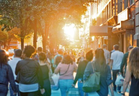 The picture shows an urban pavement alongside a densely built-up road in summer. There are several individuals visible from behind walking on the pavement, some of them carrying shopping bags. The scenery is enveloped in a warm, yellowish light from the low sun. 