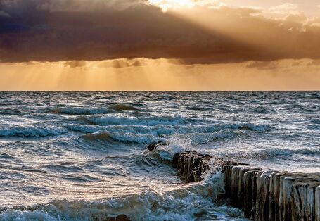 The picture shows a sectional view of a wild sea with waves crashing over projecting posts. The sky is tinged with orange and covered in dark clouds. Here and there rays of sunshine are breaking through the cloud cover.