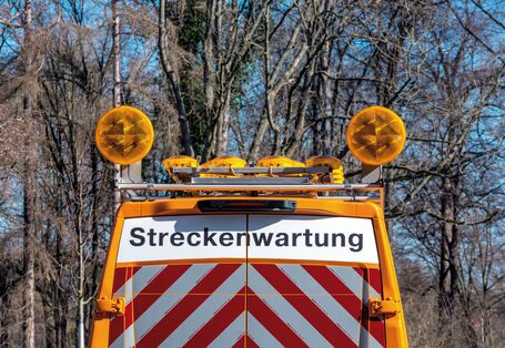 The picture shows the upper part of the tail end of a vehicle belonging to the highway maintenance service. The vehicle is orange and its rear doors are marked with the word ’Streckenwartung’ (track maintenance) and reflective red-and-white striped stickers. There are warning lights fitted to the vehicle.