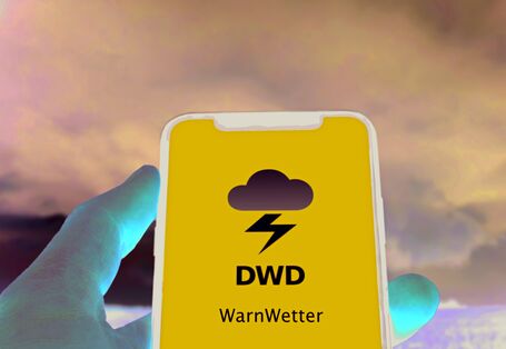 The picture shows a hand holding a smartphone. The smartphone screen is blue and shows the logo of DWD’s WarnWetter App. In the picture’s background, the black clouds of a thunderous sky are visible.