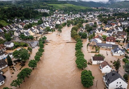 Das Bild zeigt eine Ortschaft mit Ein- und kleinen Mehrfamilienhäusern in einem Tal. Der durch die Ortschaft fließende Fluss ist über die Ufer getreten und überschwemmt die tiefer liegenden Teile der Ortschaft mit braunem Wasser. Der übliche Flusslauf ist durch Baumreihen zu erkennen. Zudem ist eine zerstörte Brücke, die den Fluss querte, sichtbar.