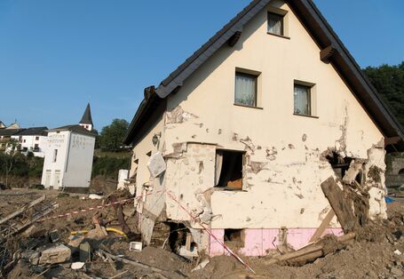 The picture shows a badly damaged single-family house. The exterior wall has been breached in several places, some windows are missing, and the exterior rendering is pitted to well above the ground floor. Mud and debris have accumulated all round the house.