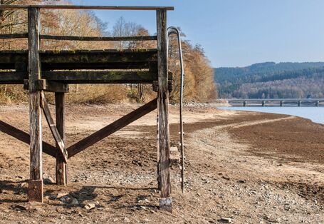 The picture shows a footbridge leading across the dried-up sandy and stony shore area of a lake. A bridge and some residual water are visible in the background.