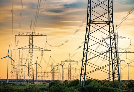 Das Bild zeigt zahlreiche Hochspannungsmasten und -leitungen vor einem wolkenverhangenen, gelblich leuchtenden Himmel. Im Hintergrund sind einige moderne Windkraftanlagen zu sehen.