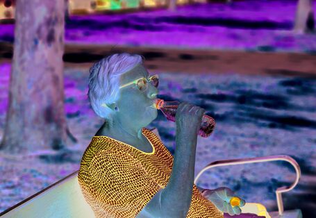 Das Bild zeigt eine ältere Frau, die auf einer Parkbank im Schatten unter einem Baum sitzt und aus einer Wasserflasche trinkt.