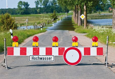 The picture shows a flooded dip in a road. There is a roadblock in the foreground indicating that the road is closed to traffic. A sign reading ‘Hochwasser’ (floodwater) is fixed to the roadblock. There are flooded meadows visible on both sides of the road.  