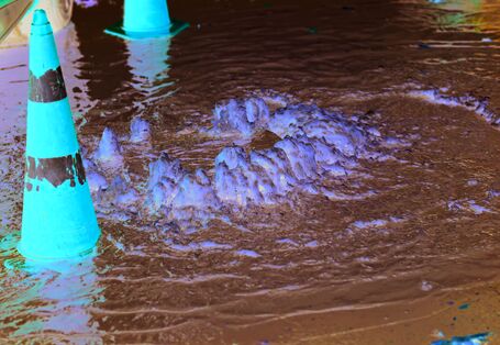 The picture shows a gully on a flooded road. The waterspout indicates where the gully is located. There are safety cones beside the gully. A car wheel is visible at the very edge of the picture.