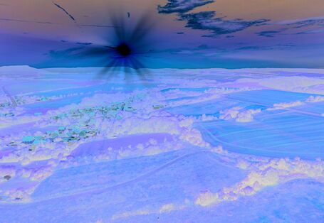 The picture shows a slightly hilly landscape characterised by agricultural activity. The centre of the picture shows a village embedded in the landscape. In an almost cloudless sky which is already tinged with orange, you can see a low sun wrapping the landscape in a warm light.