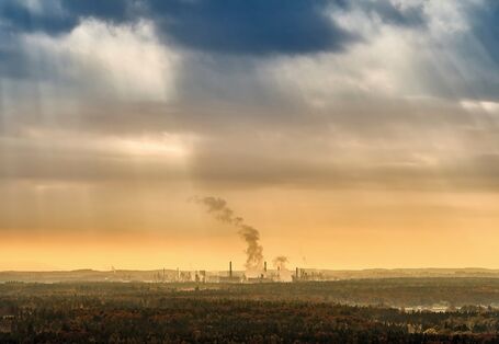 The picture shows a sectional view of a wooded landscape. There is an industrial estate visible in the distance, consisting of several buildings and facilities. There is smoke rising from some chimneys. The sky is densely cloud-covered. Above the horizon, the sky is tinged with yellow, with dark, greyish-blue clouds billowing at the margin of the picture. In a few lighter places, rays of the sun are penetrating the band of clouds.