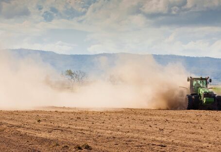 Das Bild zeigt eine trockene Ackerfläche, über die ein Traktor fährt. Aufgrund der Trockenheit wirbelt das Fahrzeug große Staubwolken auf. Im Hintergrund ist eine bewaldete Hügelkette zu erkennen.