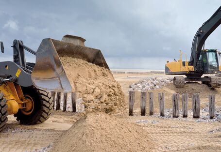 In the foreground the picture shows a full loading shovel of a wheeled loader, which is being emptied on to a sandy beach. There are wooden posts and a pile of stones visible on another sandy beach in the background. The sky is covered in dark grey clouds.