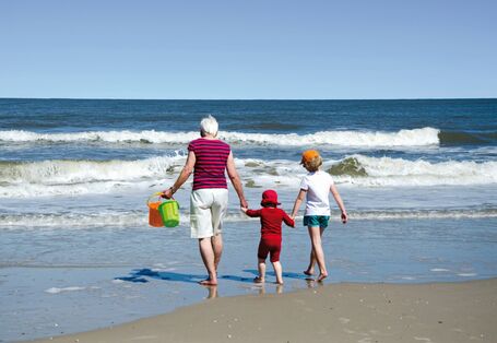 The picture shows an elderly woman, a toddler and a child of school age from behind; they are all wearing summer clothing. Holding hands, they are walking on the beach towards the waves. The woman is holding two buckets for digging in the sand.