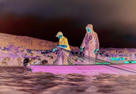 The picture shows two men in waterproof working clothes standing in a small boat on Lake Constance. One of them is holding a fishing net. He seems to be pulling it out of the water. In the background, the lakeshore and a settlement are visible.